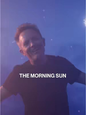A man is smiling directly at the camera in a slightly blurry image. He's set against a dark blue background that has some light-colored specks scattered around, resembling stars. Text at the bottom reads 'THE MORNING SUN'.
