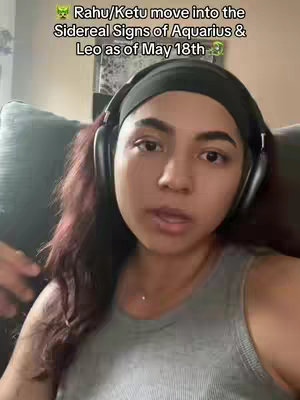 A woman with dark hair and a black headband is centered in a selfie-style video. She wears a gray tank top and has headphones around her neck. Text overlays the frame at the top, describing the current astrological events. The background is blurred, suggesting an indoor setting.
