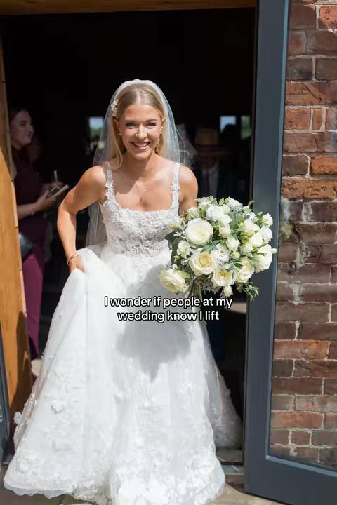A smiling bride in a white wedding dress and veil stands in a doorway holding a large bouquet. The photo is warm-toned with natural light filling the shot.