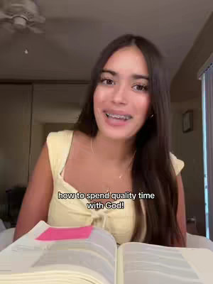 A young woman with long dark hair and braces is smiling at the camera. She is wearing a pale yellow top and has a necklace. In front of her is an open book with a pink sticky note on one page. The background is softly lit.