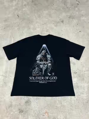 A black t-shirt is shown on a gray surface, with a design on the front featuring a kneeling soldier in armor, holding a sword. The design is set against a bright, ethereal light.  Below the image is the text 'SOLDIER OF GOD' and a bible verse.