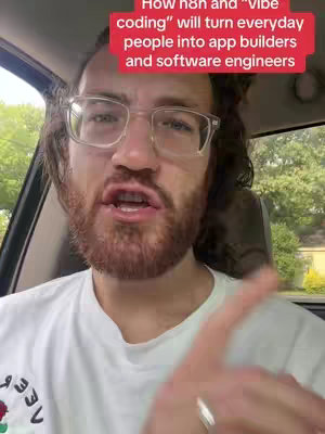 A selfie-style shot from inside a car. A man with glasses, a beard, and a white t-shirt is speaking, with a red banner above him containing white text. The background shows a blurred view of a road with trees and buildings.