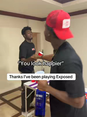 Two young men are in a room. One is wearing a red cap, and the other a black t-shirt. The cap-wearer is facing the camera, while the other is slightly angled. Text overlays read, 'You look happier' and 'Thanks I've been playing Exposed'.
