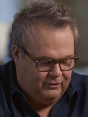 A close-up shot of a man wearing glasses. The background is blurred, and the man is looking down.
