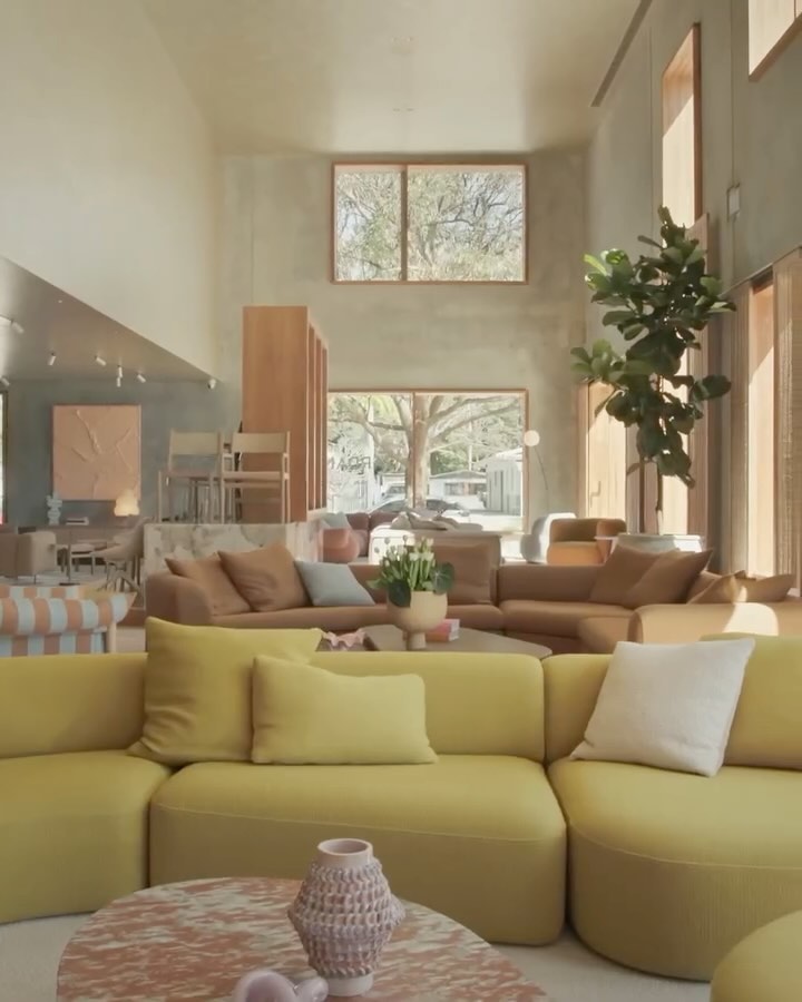 The image showcases the interior of a modern, open-plan showroom filled with furniture. Dominant colors are mustard yellow, soft browns, and neutral grays. A large yellow sectional sofa is prominently placed in the foreground, with accent pillows. A round marble coffee table sits in front of the sofa. To the right, a tall fiddle-leaf fig tree adds a touch of nature. In the background, a series of windows bring in natural light, and the overall design conveys a sense of spaciousness and elegance. The lighting is bright and natural, highlighting the textures and tones of the materials used in the showroom.