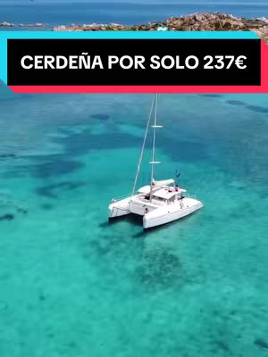 An aerial view of a picturesque coastal scene. The turquoise water is crystal clear, revealing the sandy bottom below. A white catamaran is sailing in the water, creating a sense of serenity. The background features a coastline with lush greenery and hills under a clear sky.
