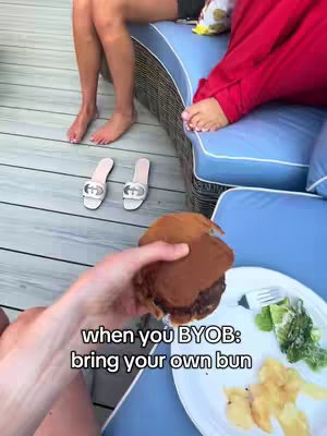The image is a first-person perspective shot, likely taken outdoors, of someone holding a gluten-free bun with a burger patty inside. A plate with a side of salad and fries sits to the right. In the background, two people are sitting on a blue and wicker outdoor sofa, with feet visible in the foreground, beside Gucci sandals on wooden flooring.