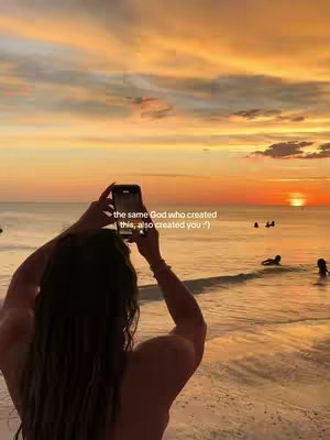 A woman stands on a beach taking a photo of a sunset with her phone. The sky is filled with orange and yellow hues, and the ocean is calm. There are a few people in the distance in the water. The text overlay reads: "the same God who created this, also created you :) "