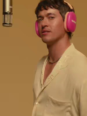 A man wearing a cream-colored shirt and pink headphones stands in front of a tan background. The man is looking slightly to the side, and a microphone is partially visible in the upper left corner.