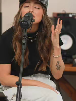 A woman with long brown hair, wearing a black t-shirt and a cap, is singing into a microphone. She has her eyes closed and her hand raised, appearing to be in a recording studio. There is a tattoo on her forearm. Studio equipment is visible in the background.
