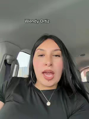 A woman is seated in a car, facing the camera directly. The top half of her body is visible. She has shoulder-length, black hair, and is wearing a black top and a silver necklace. Her expression is one of open-mouthed speech. The interior of the car is visible in the background.