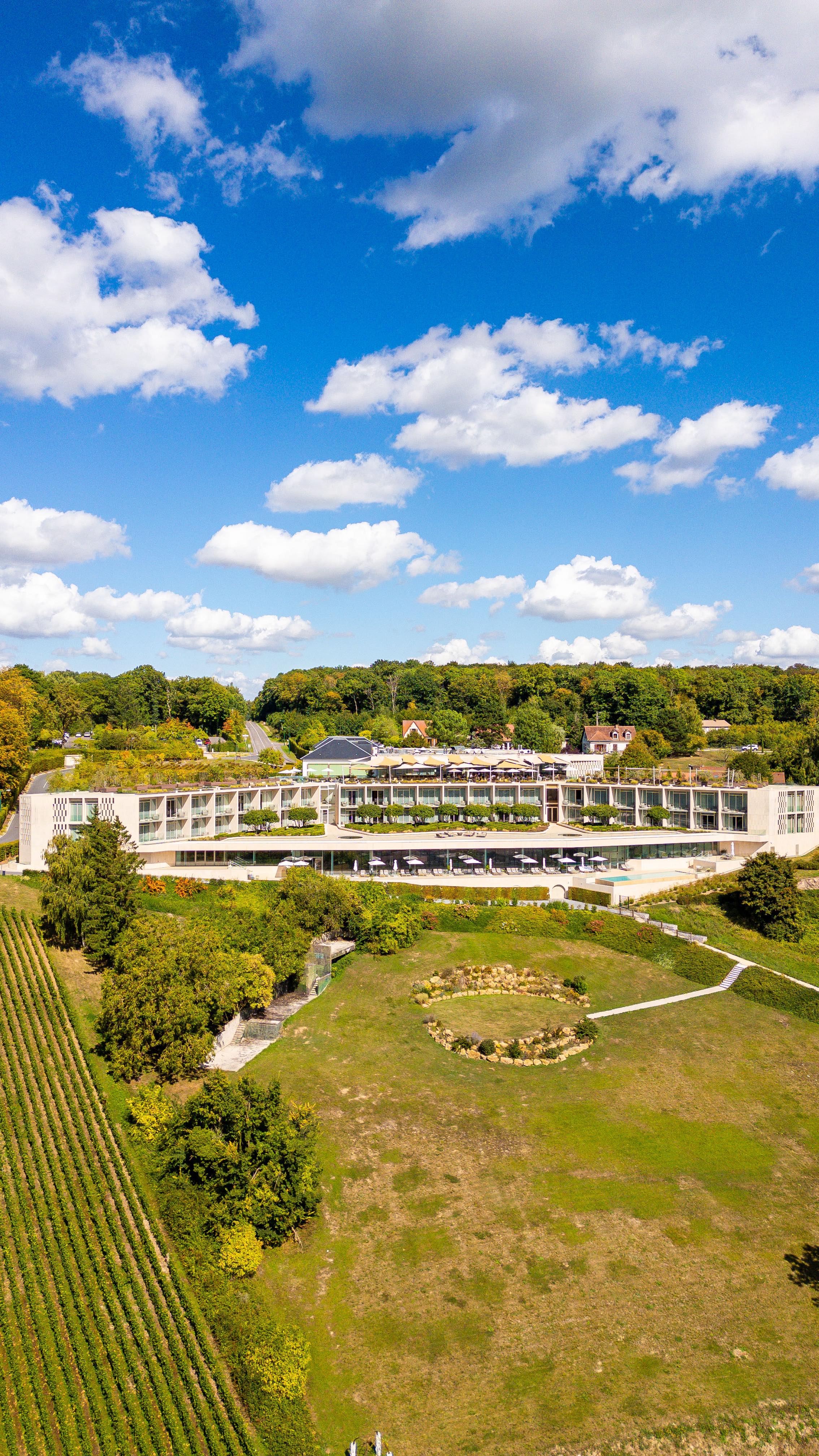 An aerial view of a luxury hotel and spa, set amidst rolling green hills and vineyards under a blue sky with fluffy white clouds. The hotel has a modern design with a light-colored facade, green rooftops, and large windows. Lush green lawns stretch out below the hotel, leading to neatly planted rows of grapevines. 