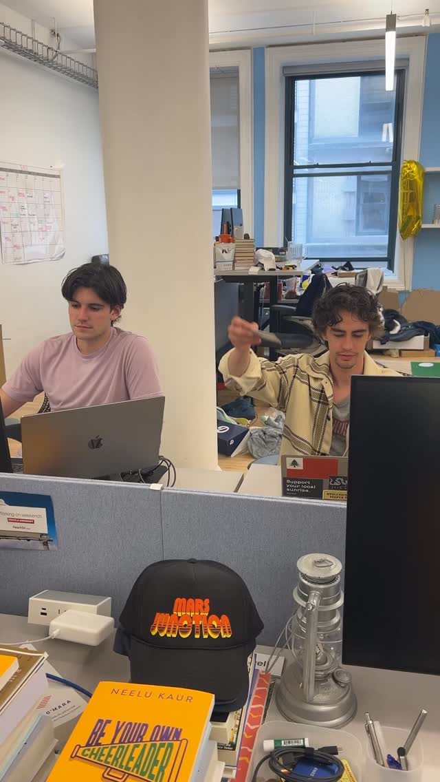 An office setting with two men working at individual workstations. The foreground features a desk with various items, including a cap, books, and office supplies. One man is looking towards the screen while the other is holding something in the air near his head. The overall tone is casual and professional.