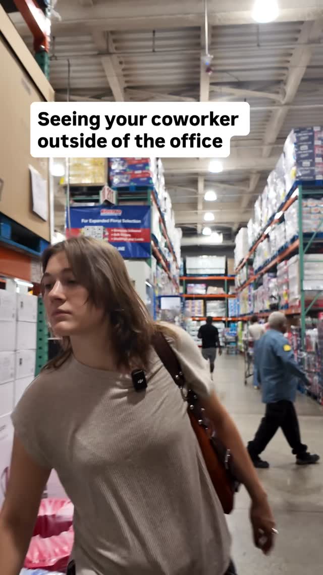 A woman is walking in a large warehouse store, looking to her left, with shopping carts and product displays in the background. The scene is brightly lit, suggesting a daytime setting.