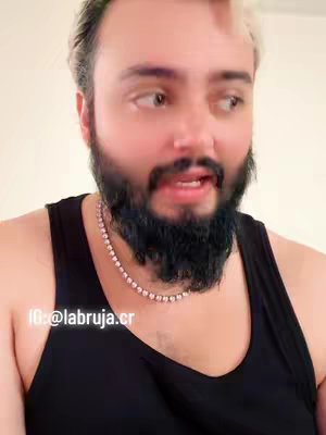 A medium shot features a light-skinned man of mixed ethnicity looking slightly to his right. His hair is short and blonde on top, with hints of darker roots and his face is framed by a meticulously styled black beard. His eyes are open and he appears to be speaking. He is wearing a black tank top, and a silver chain necklace is visible at his neck. The background is a plain, off-white wall. The bottom left corner of the frame features white text with the creator's ID. The overall presentation suggests a personal, communicative tone.