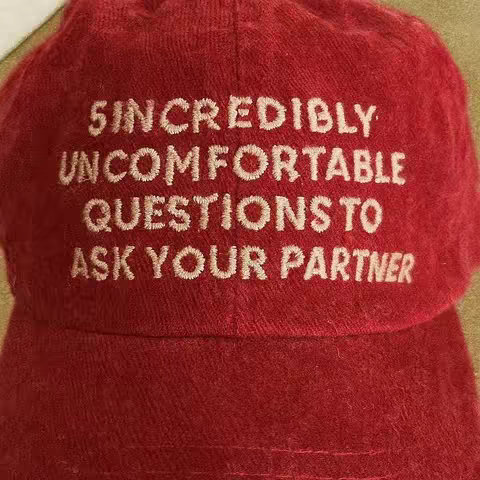 The image shows a close-up of a red cap with white embroidered text. The cap is a deep, slightly faded red, and the texture suggests a soft material like velvet or corduroy. The text, in a stylized serif font, reads in multiple lines, "5 INCREDIBLY UNCOMFORTABLE QUESTIONS TO ASK YOUR PARTNER". The lighting appears soft and diffused, possibly from indoor conditions, highlighting the texture and the embroidery. The composition is simple, with the slightly angled cap filling most of the frame.