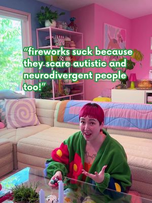 The image shows a person with short, bright pink hair looking at the camera.  They're wearing a green robe with floral accents and gesturing with their hands.  Text in a speech bubble reads, “fireworks suck because they scare autistic and neurodivergent people too!” The background setting includes a couch and a window, the room is brightly coloured with pink and blue walls and plants.
