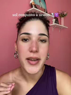 A close-up shot of a woman speaking, set against a softly lit pink background. She has dark, curly hair and is wearing a sleeveless, dark purple top. Her expression is open and engaged as she appears to be talking. On the upper part of the background are some white shelves with decorations with a small figurine.
