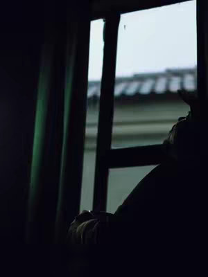 A dimly lit indoor shot, possibly a room, with heavy shadows. A window is visible on the right side of the frame, and a portion of an exterior, likely the roof of a building, is seen through it. The main subject is silhouetted, with a person's arm and hand holding a dark object, such as a cup or a phone, visible. The image is dominated by cool colors with emphasis on darkness and light from outside the window.
