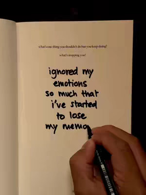 A hand holding a black pen writing on an open book. The book is on a beige surface and has text already written in a handwritten font. The top of the image shows text that reads, "what's one thing you shouldn't do but you keep doing?\nwhat's stopping you!", above the handwritten text which reads, "ignored my \nemotions\nso much that\ni've started \nto lose\nmy memo".