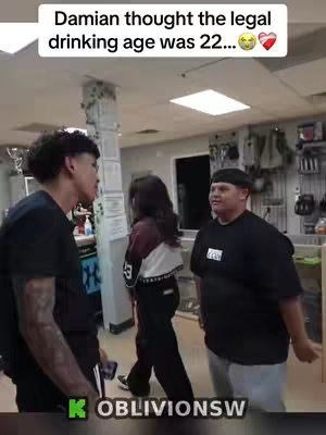 The image shows three individuals in an indoor setting. On the left, a young man with short, dark, curly hair is facing right, appearing to be speaking. Behind him, a woman with dark hair is walking away facing left. On the opposite side, a man with a black cap stands casually. Above them, a text overlay says 'Damian thought the legal drinking age was 22...'. The lighting is bright with general indoor illumination.