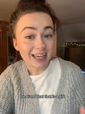 A young woman with fair skin and light brown hair pulled back in a bun is captured in a selfie-style shot. She wears a light gray knit sweater over a white t-shirt. Her expression is lively, with her mouth open as if speaking, and her eyes sparkling. The background shows a dimly lit interior with glimpses of a wooden cabinet and a window partially visible, suggesting a home setting. A text overlay at the bottom of the screen repeats a key phrase from the video's message, reinforcing the video's core theme. Overall, the aesthetic is warm and inviting.