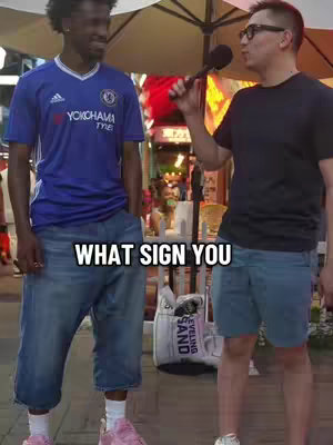 Two men are standing outside, one with a microphone interviewing the other. The man being interviewed is wearing a blue Chelsea soccer jersey and jean shorts. The interviewer is wearing glasses with a dark shirt and blue shorts. The text overlay says, "WHAT SIGN YOU".