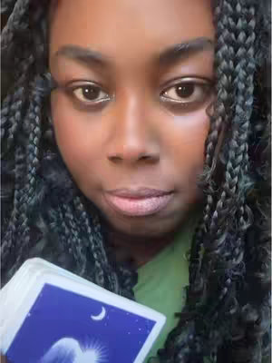 Close-up shot of a Black woman looking directly at the camera. She has braided black hair, and is wearing a green shirt. She is holding a deck of tarot cards in her hands.