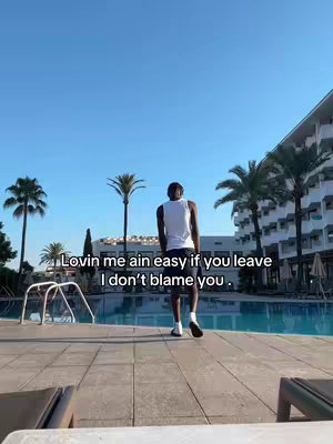 A man in a white tank top and dark shorts walks away from the viewer along a tiled surface beside a pool. Palm trees and a building with many balconies form the background. The sky is a clear blue.