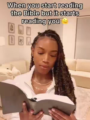 A woman with long, braided hair sits indoors, holding a book open in her hands. She's wearing a white top and a gold necklace, with a neutral-toned room and furniture in the background.