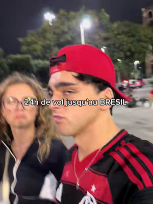 A selfie-style shot featuring a young man in a red cap and a Flamengo soccer jersey, standing with a woman on a street at night. The foreground features the man with slightly blurred features and a woman to his left. The background includes indistinct lights and buildings. Text overlayed on the image reads, \"24h de vol jusqu'au BRÉSIL\".