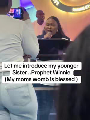 A woman in a formal setting speaks into a microphone. She is on a stage in front of a backdrop with a large gold design, potentially a church. A person is filming the scene from the front, while others watch. The text overlay indicates the speaker is introducing someone.