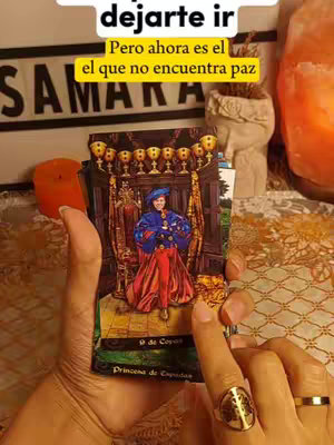 A person's hands holding a tarot card depicting a figure in regal clothing. The scene includes a candle, decorative objects, and a gold ring, suggesting a spiritual or metaphysical context.