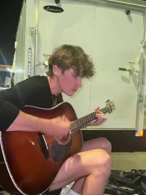 A young man with curly brown hair is playing a brown acoustic guitar. He is seated, with his legs together, in front of a silver truck and a white, metal wall. His shirt is black and the guitar is positioned in front of him.