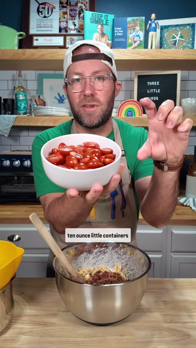 A man in a green shirt and glasses is holding a white bowl filled with halved red tomatoes. He is standing in a kitchen with various items in the background, including shelves with books and decorations, and a mixing bowl on the counter in front of him.