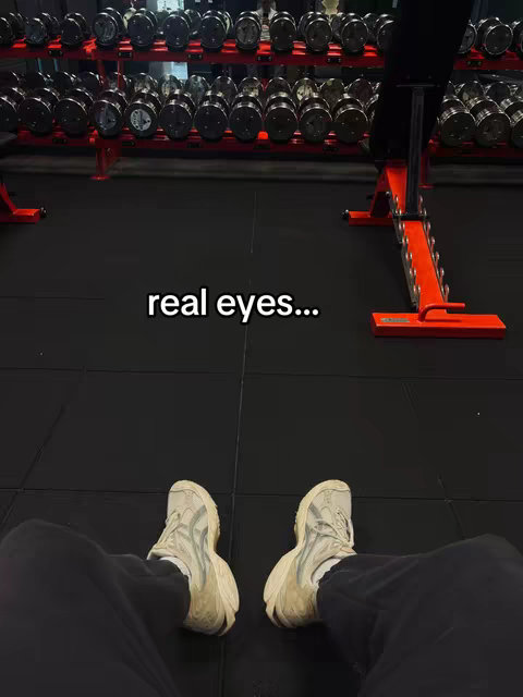 The image shows a gym setting. The camera is positioned looking down at the user's feet, showing the user's shoes and portions of their legs. Behind the person's feet, there is gym equipment visible, including dumbbell racks, a weight bench, with black flooring throughout.
