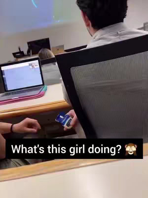 The image shows a classroom with a laptop open on a desk. The main focus is on a hand holding a tube of what appears to be Vaseline. Another person is shown looking at the laptop, and a blurred image is visible against a screen. The text overlay asks what the girl is doing.