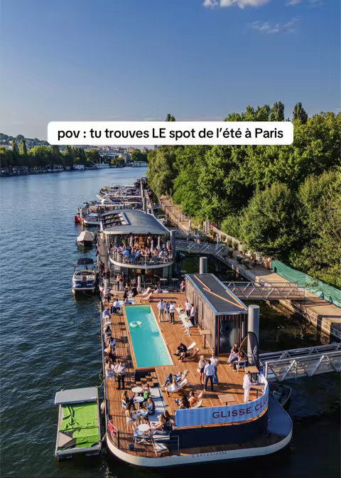 An aerial view of a barge on a river, with various sections for dining, lounging, and a small pool. People are enjoying the sunny day. The background shows trees, a bridge, and buildings.