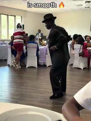 An image from inside a venue or event shows a man in a dark suit, black hat, and black shoes, appearing to dance or pose mid-step.  Other people are in the background including a person wearing red and white striped shirt.