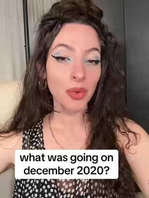 A woman with long, dark hair is centered in the frame, facing the camera. She has noticeable winged eyeliner with aqua-colored eyeshadow, and her lips are a bold pink. She wears a black and white spotted top and a gold necklace and earrings. A white rectangular text box is placed in the lower center of the image with the text in all lowercase letters: "what was going on december 2020?"