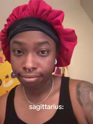 A close-up shot of a person with dark skin. They are wearing a black tank top, a silver necklace, and a red bonnet. The person has a nose ring and expression, with text below displaying "sagittarius:"