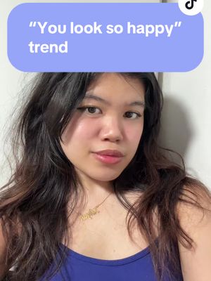 A young woman looks directly at the camera. A purple bubble with white text that says \"You look so happy\" trend overlaps her face. She has dark, wavy hair, dark eyeliner, and a necklace that has a word in cursive on it. A blue tank top is visible.