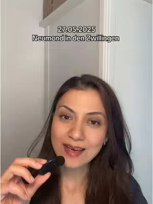 A woman with dark hair is smiling and holding a microphone in front of her. Text overlay at the top reads '27.05.2025 Neumond in den Zwillingen'. The background is a plain white wall.
