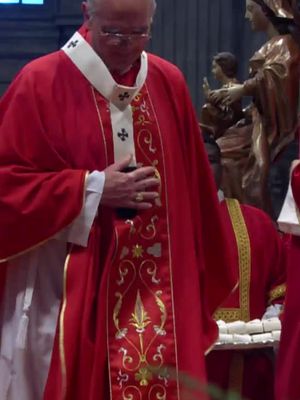 A man in a red religious garment is the central figure, seen from the chest up. He has a white collar and is wearing eyeglasses. The garment has gold embroidered patterns down the front. The background is dark with a glimpse of a figure. The lighting is dim.