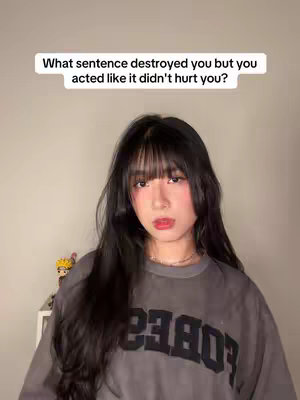 A young woman with long black hair and bangs looks directly at the camera. Above her, a text overlay poses the question "What sentence destroyed you but you acted like it didn't hurt you?" She wears a gray t-shirt with a black logo. The background is a plain, light-colored wall, with a small shelf visible on the left with a small Naruto figure. The lighting is soft and even.