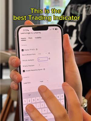 A person's hand is holding a smartphone displaying the 'Inputs' settings of a trading indicator app. The screen shows technical details such as 'Risk to Reward Ratio' and 'TP & SL Multiplier'. Above the phone text that reads 'This is the best Trading Indicator'. The setting's background and the person’s clothes are not clearly visible.