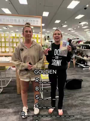 Two young women stand side-by-side in a retail environment, in front of a display. The woman on the left is wearing a tan hoodie and white shorts, holding a black object while the one on the right is holding a book and a board game. The game is partly visible. Below the women is a word puzzle with the word GOLDEN revealed and some of the letters on the bottom row. Overhead lighting is bright within the retail space.