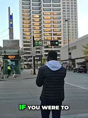 A man stands on a city street corner, his back to the camera. He is wearing a black puffer jacket and a gray hoodie. The background features a tall, modern building with many windows reflecting light, a bus stop, and traffic lights.