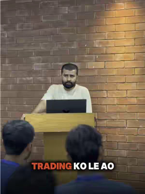 The image features a man standing behind a wooden podium, facing three people whose backs are towards the camera. The background is a brick wall, and text at the bottom of the image reads "TRADING KO LE AO". The man is wearing a cream-colored shirt and appears to be giving a presentation or lecture.