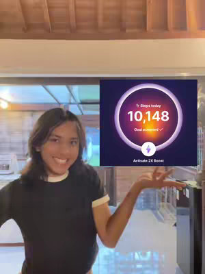 A young person with long hair is smiling while holding their left hand open, near a phone which shows that they completed a fitness challenge The background is a room with natural light.